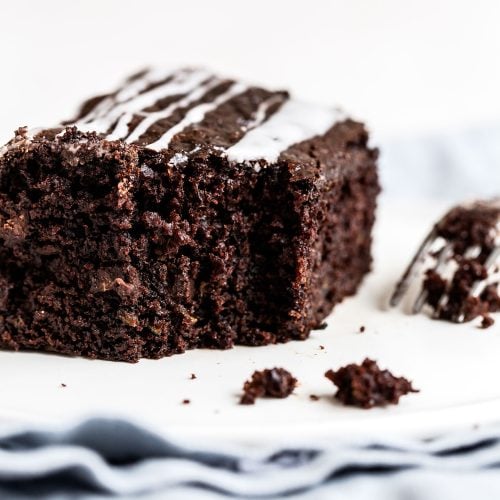 Chocolate Zucchini Cake slice with white icing drizzled on top, sitting on a white plate